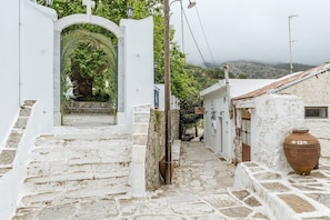 A narrow stone pathway leading to a white-walled building with a large ceramic pot on the right.
