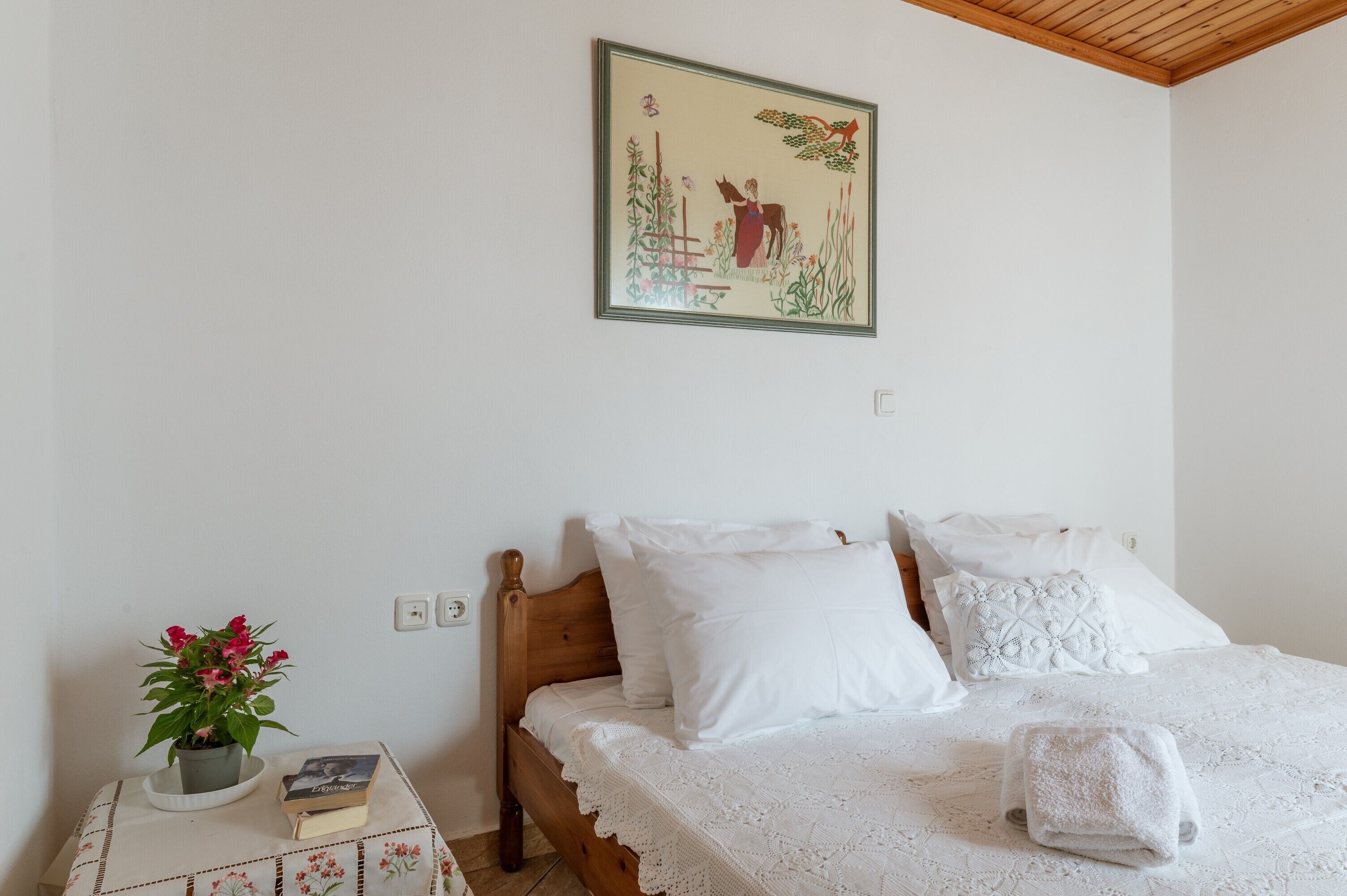 Une chambre à coucher avec un lit en bois, du linge de lit blanc, une petite table de chevet sur laquelle se trouvent un livre et une plante en pot, et un tableau encadré accroché au mur.