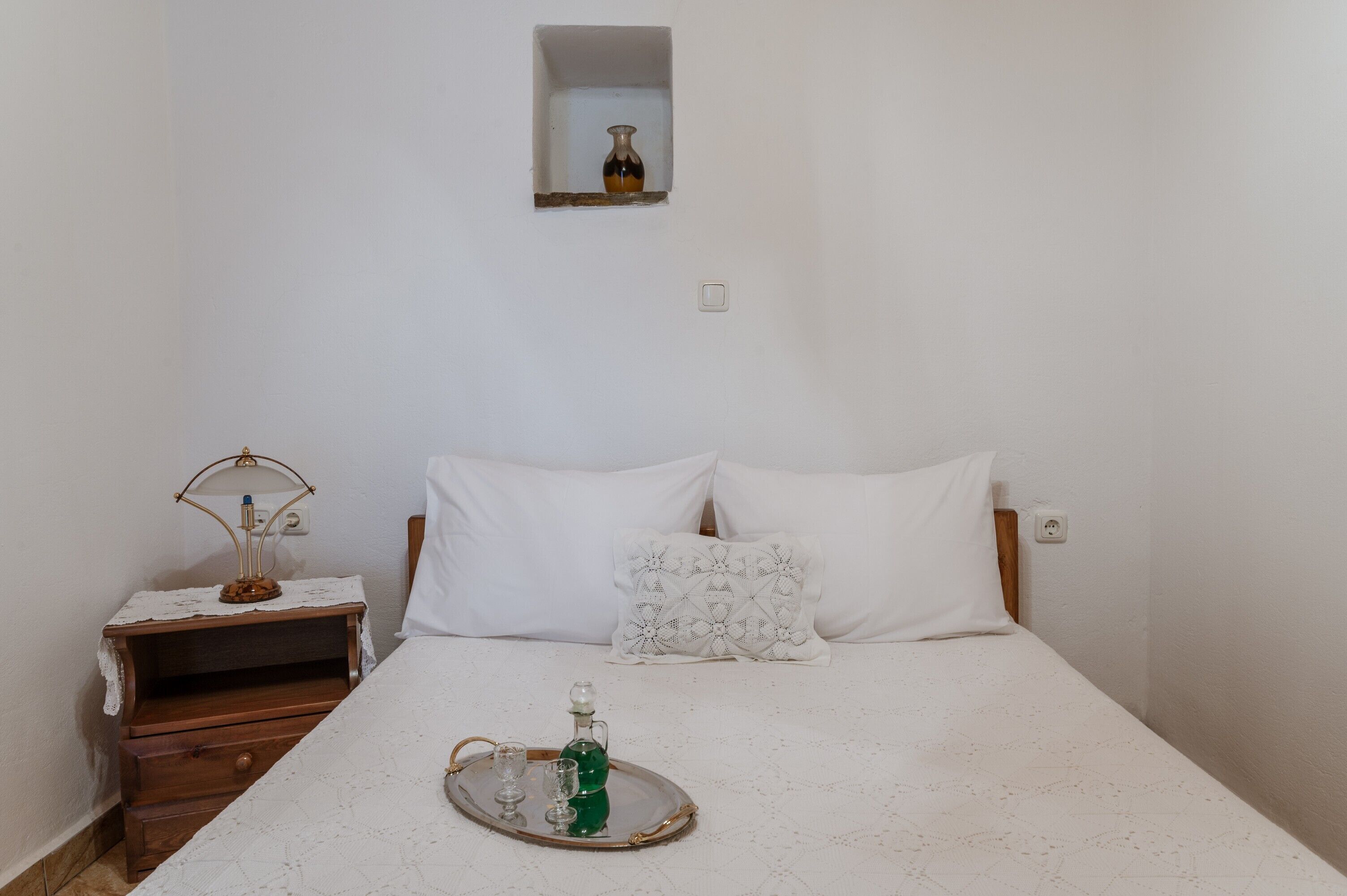 Een slaapkamer met een bed, een nachtkastje, een lamp, een dienblad met glazen en een fles, en een kleine spiegel aan de muur.