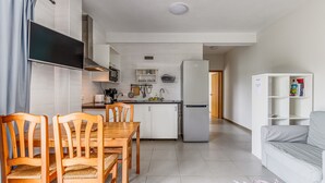 Une cuisine moderne avec des armoires blanches, un four encastré et un réfrigérateur. On y trouve un coin repas avec des chaises et une table en bois, ainsi qu’un canapé blanc dans le salon.