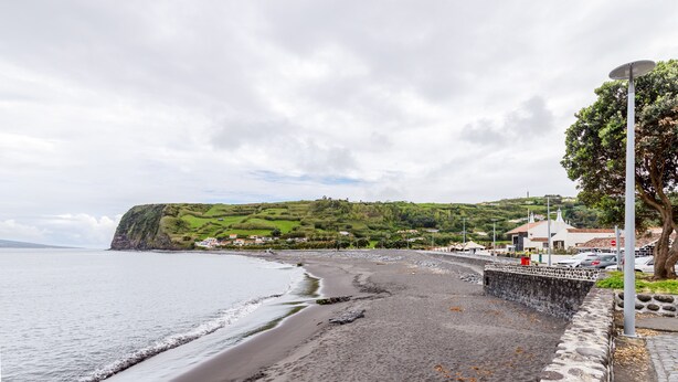 Une plage côtière bordée d’un mur de pierres, avec une promenade pavée et une vue sur une colline verdoyante où l’on aperçoit des bâtiments au loin.