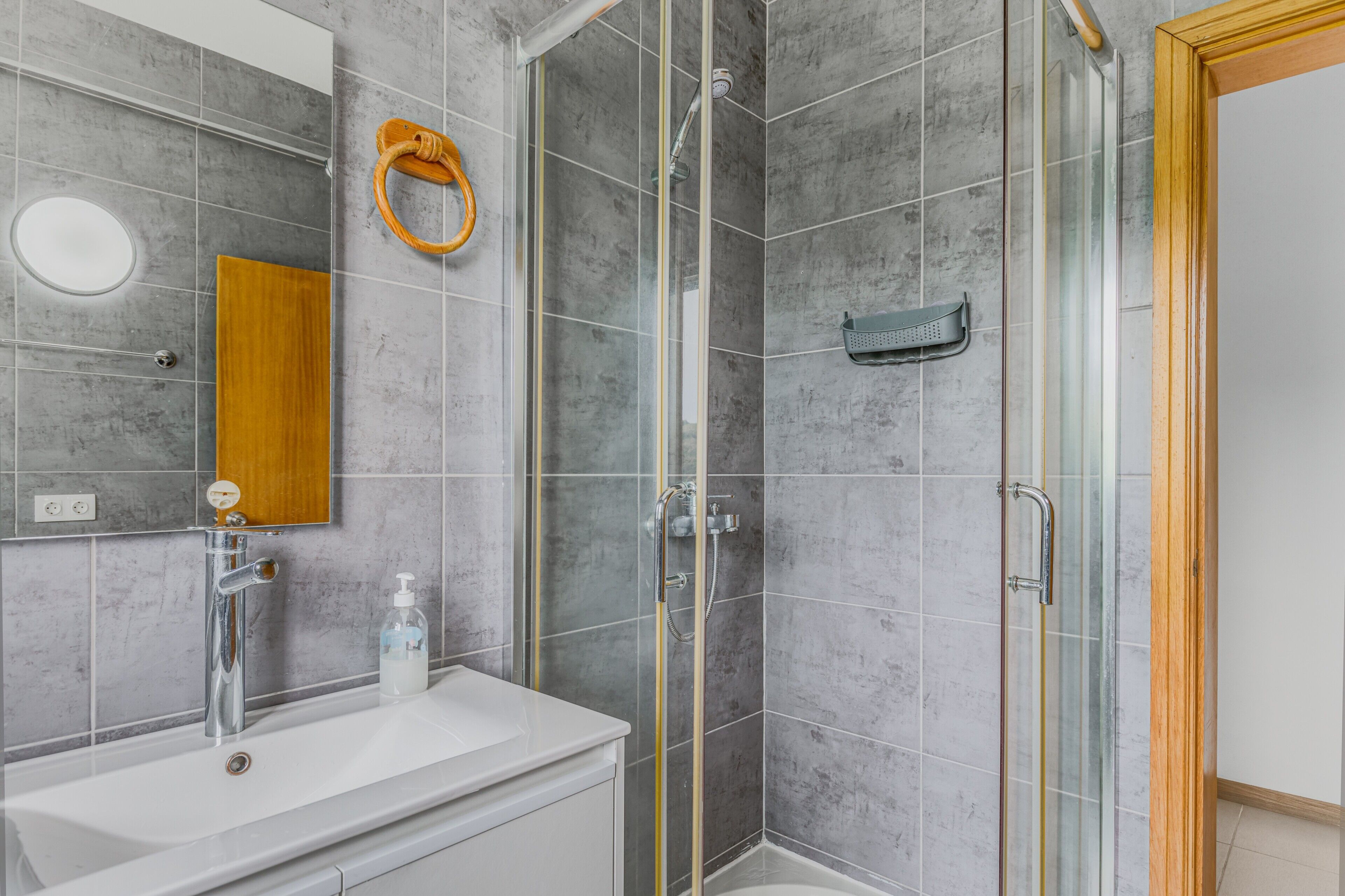 Un baño con ducha vidriada, espejo con marco de madera y lavamanos blanco con dispensador de jabón.