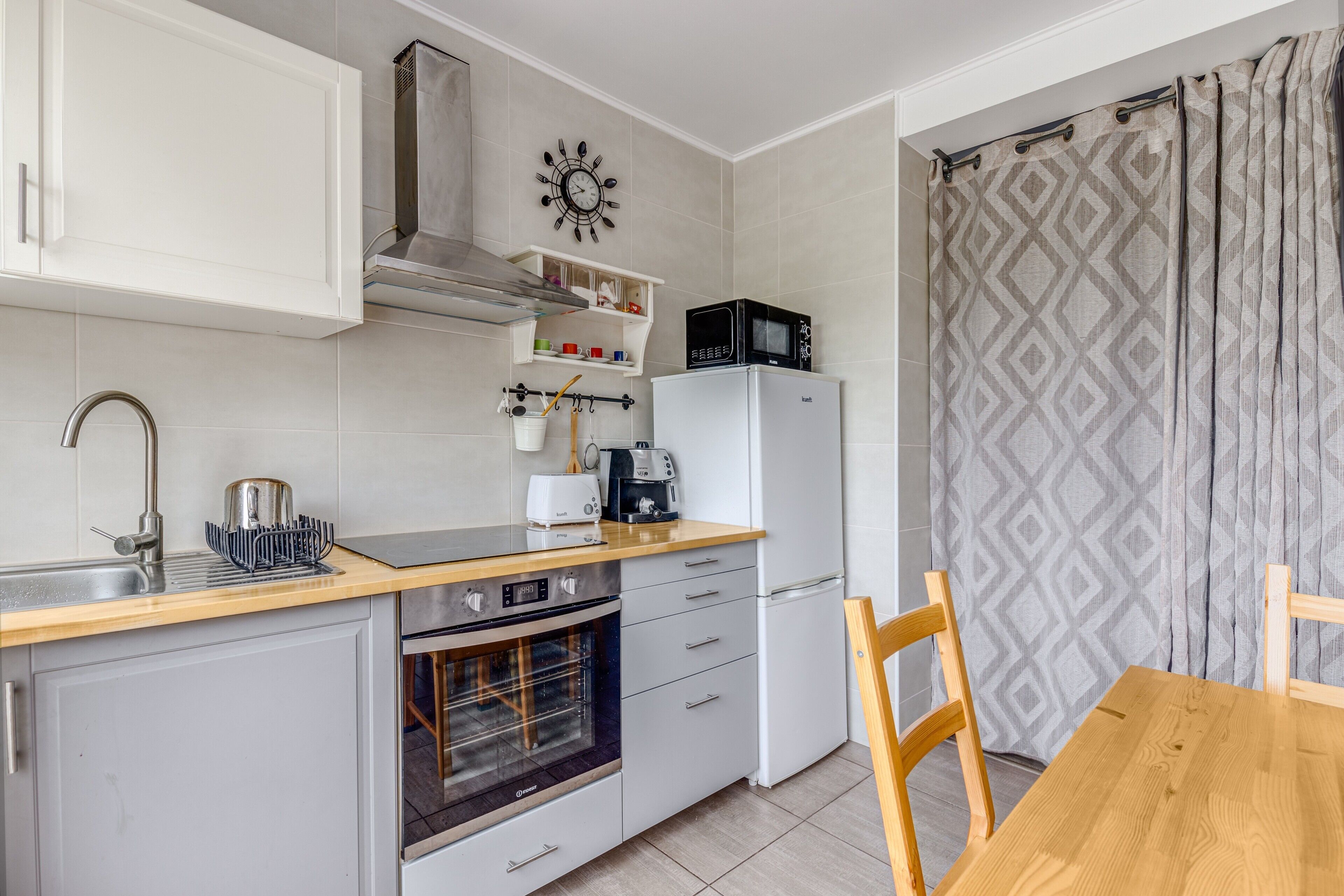 A compact kitchen with a microwave, toaster, and oven, a wooden dining table, and a clock on the wall.