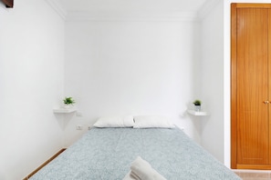 Une petite pièce avec un lit, deux étagères fixées au mur où sont posées des plantes, une porte en bois et un mur blanc.
