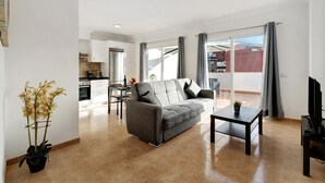 Una sala de estar con sofá, una mesa de centro y un mueble para la televisión. Se ve una cocina al fondo con armarios y una zona de comedor.