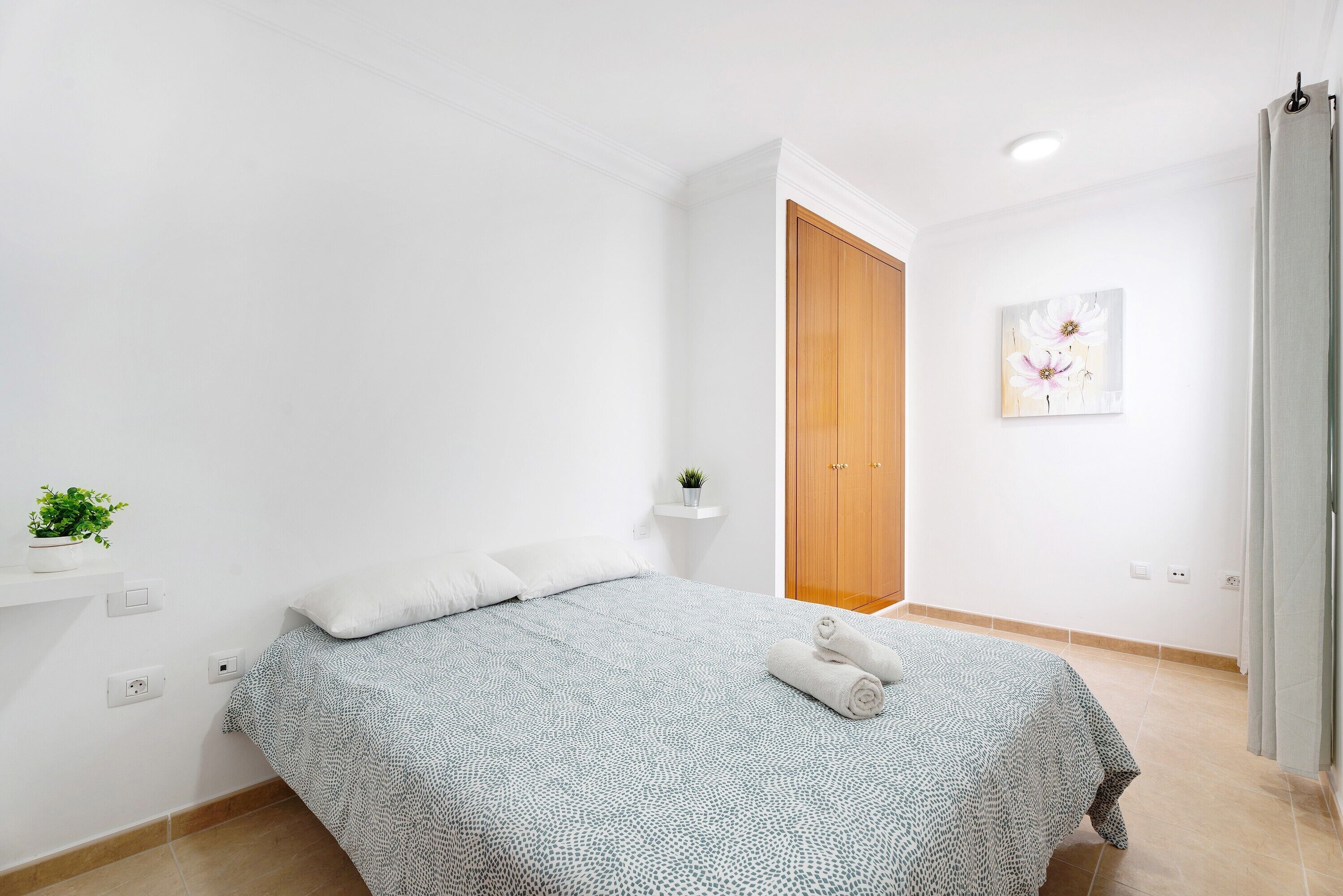 Un dormitorio con cama, un armario de madera, una pequeña estantería con una planta y un cuadro en la pared.