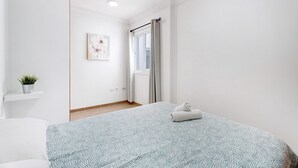 Un dormitorio con una cama, una ventana con cortinas, una planta pequeña en un estante y un cuadro en la pared.