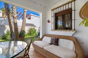 A wicker sofa with cushions, a glass table, and chairs in a sunlit room with a view of palm trees and a building. - Villa 'Casa Dos Mares' with Private Pool, Private Terrace and Wi-Fi (Cádiz)