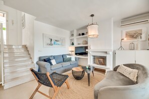 A modern living room with a staircase, a grey sofa, a wicker chair, a round wooden table, and a grey armchair.