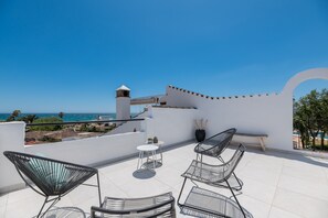 A rooftop terrace with a view of the sea, featuring a white wall, a chimney, and black metal chairs.