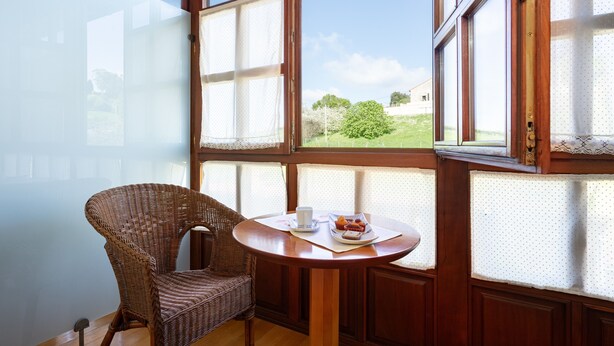 Een kamer met een rieten stoel, een ronde houten tafel en grote ramen met uitzicht op groen.