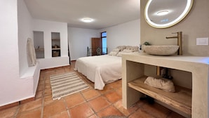 A modern bedroom with a large bed, a vanity with a round mirror, and a view of the outdoors.