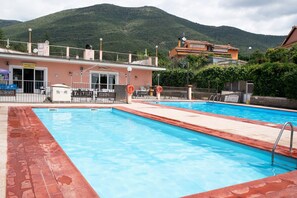 Una piscina con montañas al fondo, un edificio con un letrero y un área cercada con mesas al aire libre.