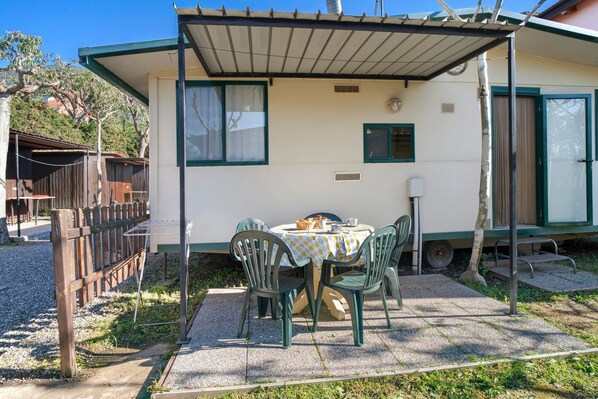 Une terrasse couverte avec une table et des chaises, un petit bâtiment doté d’une fenêtre et une clôture en bois.