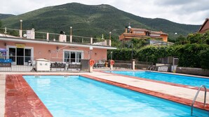 A swimming pool with a mountain backdrop, a building with a sign, and a fenced area with outdoor seating. - Mobile Home "Casa Mobile Lux La Pineta 21" with Shared Pool, Private Terrace & Wi-Fi (Albenga)