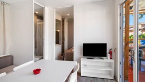 A hotel room with a white table, a TV on a white stand, a red object on the table, and a view of the outside through a glass door.