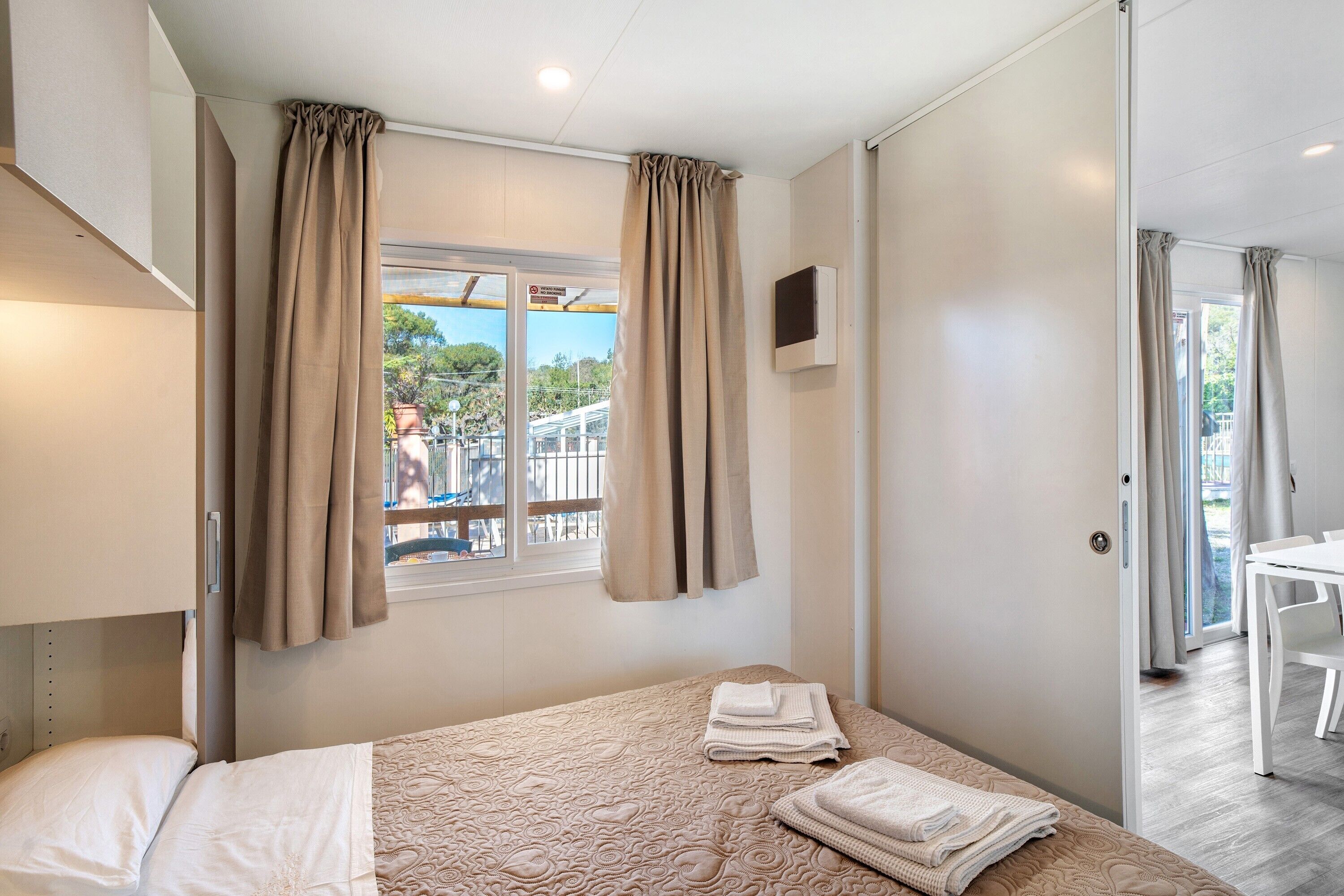Ein Hotelzimmer mit Bett, Vorhängen am Fenster, einem kleinen Tisch und Blick ins Freie.