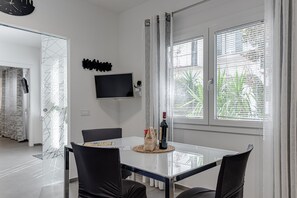 A modern dining area with a glass table, black chairs, a bottle of wine, and a TV on the wall.