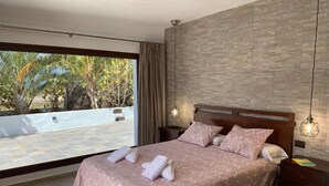 Un dormitorio con una cama grande, una mesita de noche, una lámpara colgante y vistas a una terraza con palmeras.