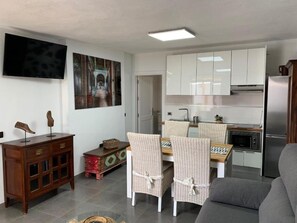 A living room with a sofa, a dining table with chairs, a kitchen area with cabinets and appliances, and a television on the wall. - Apartment 'Finca Wapa - Margarita' with Sea View, Wi-Fi and Air Conditioning (Frontera)
