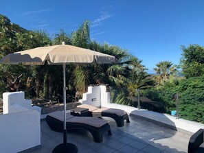 A rooftop terrace with lounge chairs, a large umbrella, and lush greenery in the background.