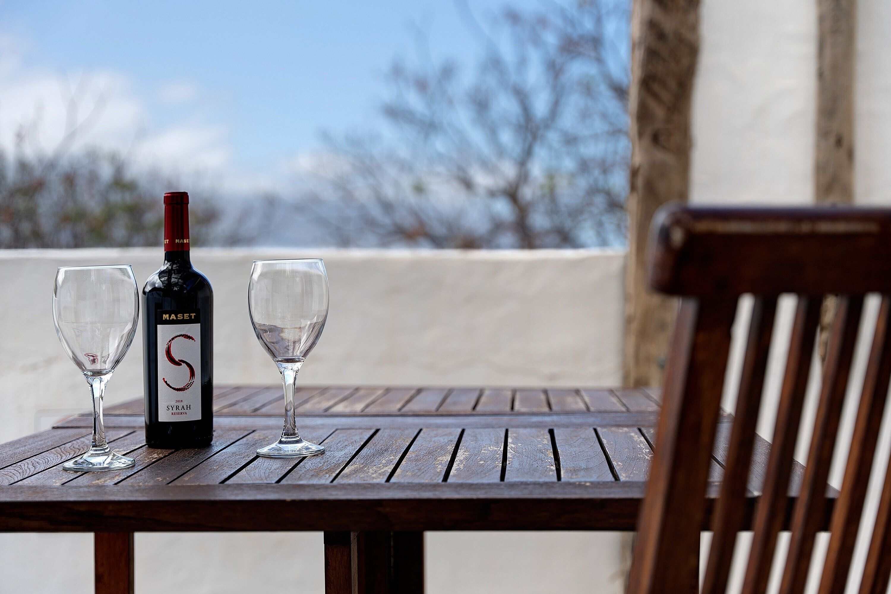 A wooden table with a bottle of wine and two glasses on it, set outdoors with a view of trees in the background.