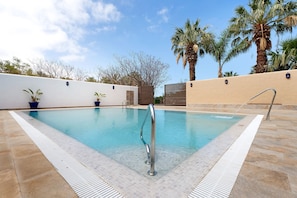 Una piscina con una escalera de metal, terraza con azulejos y palmeras al fondo.