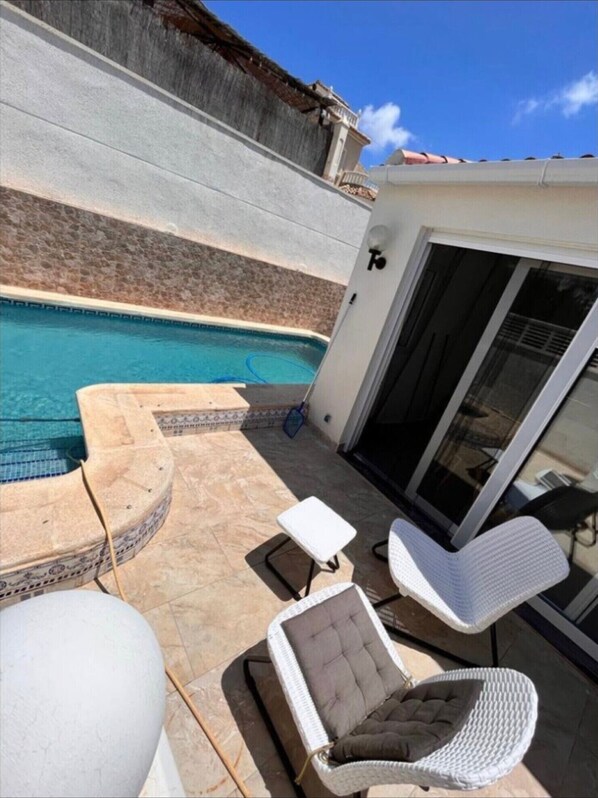 A poolside area with a pool, a chair, a small table, and a glass door leading to a covered patio.