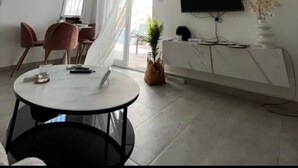 Ein modernes Wohnzimmer mit Deckenventilator, einem Essbereich mit rosafarbenen Stühlen, einem an der Wand montierten Flachbildfernseher und einer weißen Kommode mit Pflanze.