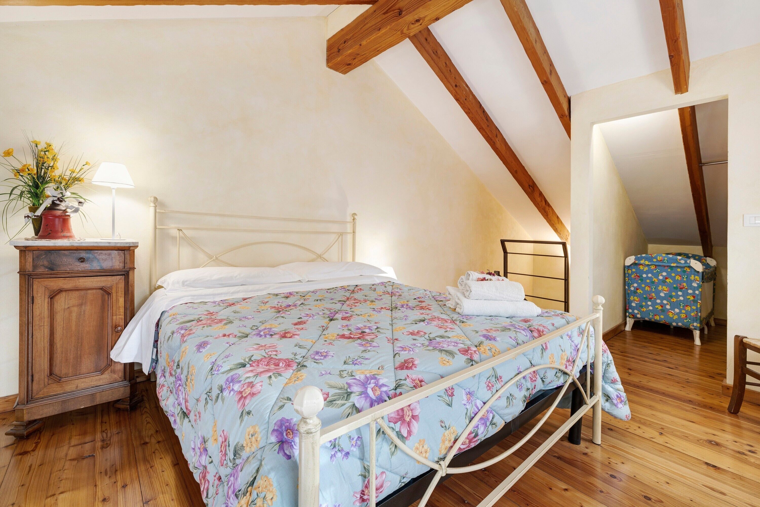 Un dormitorio con cama, piso de madera, una mesita de noche, una lámpara, un aparador y una pequeña mesa con una silla.