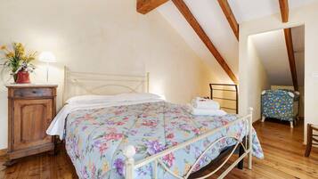 Un dormitorio con cama, piso de madera, una mesita de noche, una lámpara, un aparador y una pequeña mesa con una silla.