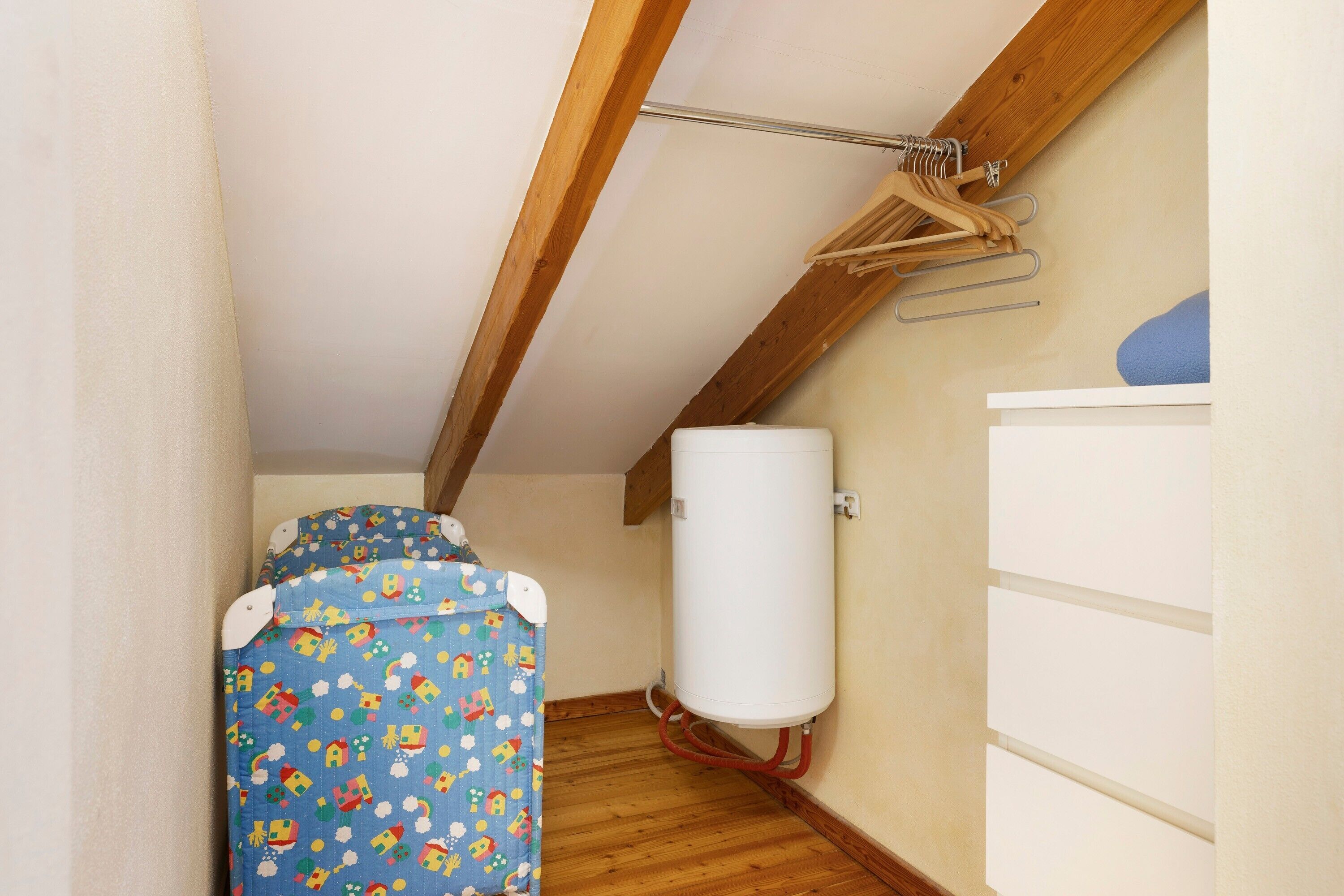 Una stanza modesta con soffitto in legno, un termosifone bianco, un letto azzurro con decorazioni e un armadietto bianco.