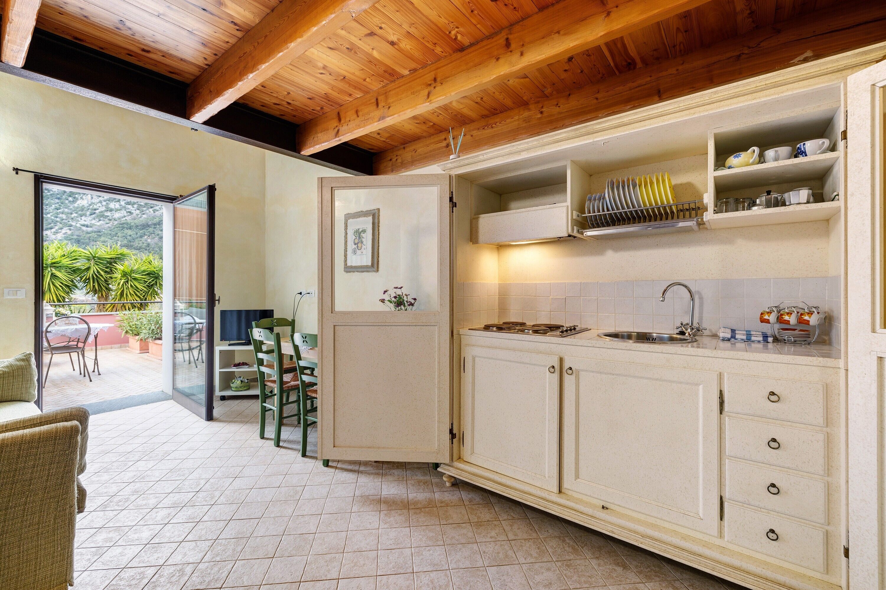 Una cocina con vigas de madera, una puerta corrediza de vidrio que da a un patio y un estante empotrado con vajilla.