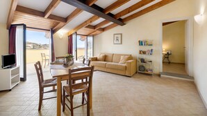 Ein geräumiger Raum mit hohen Decken, Holzbalken, einem Essbereich mit Tisch und Stühlen, einem Sofa, einem Bücherregal und einem Fernseher.