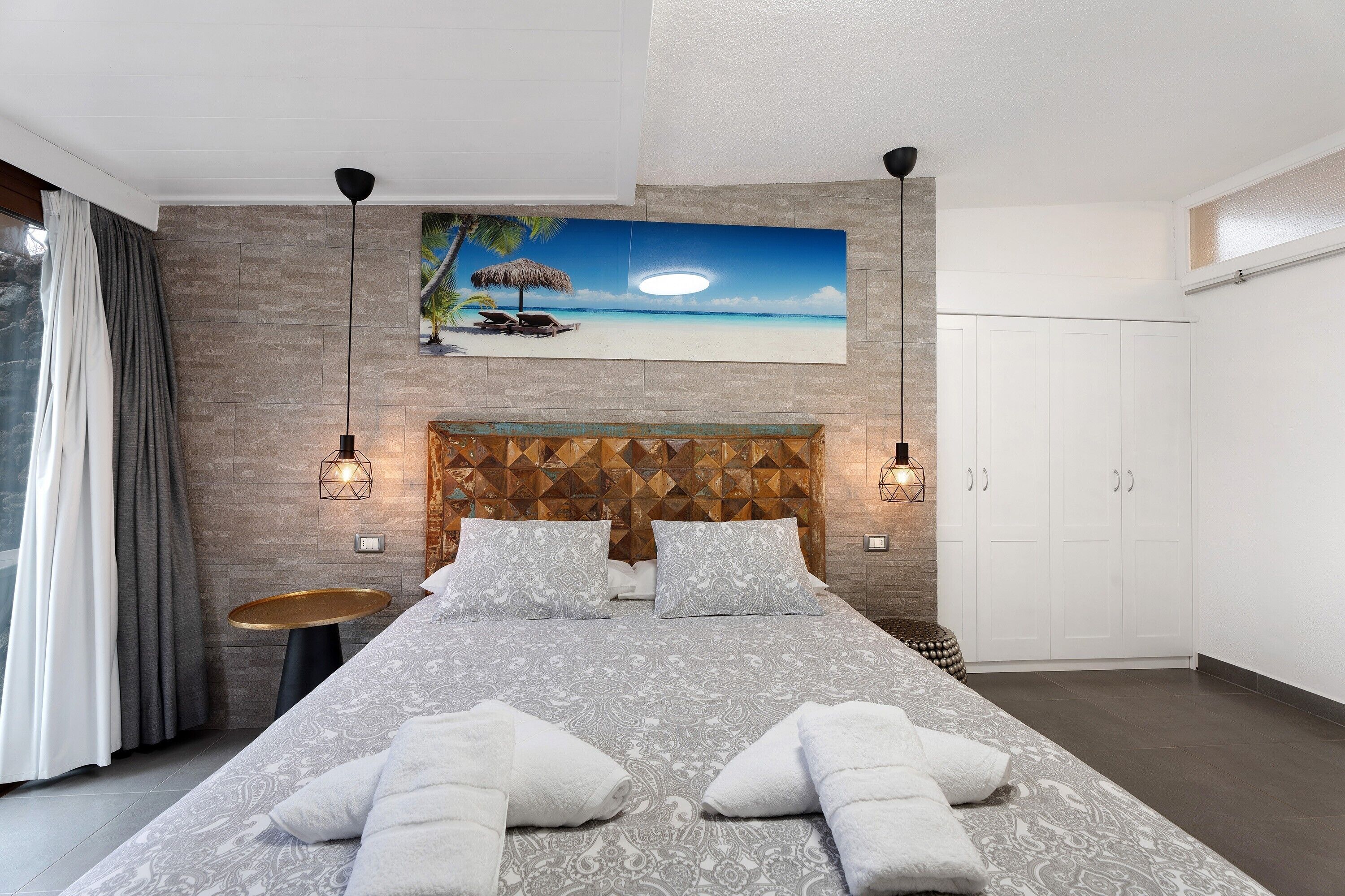 Een moderne slaapkamer met een bed, een schilderij van een strandgezicht, een rond bijzettafeltje en een kledingkast.