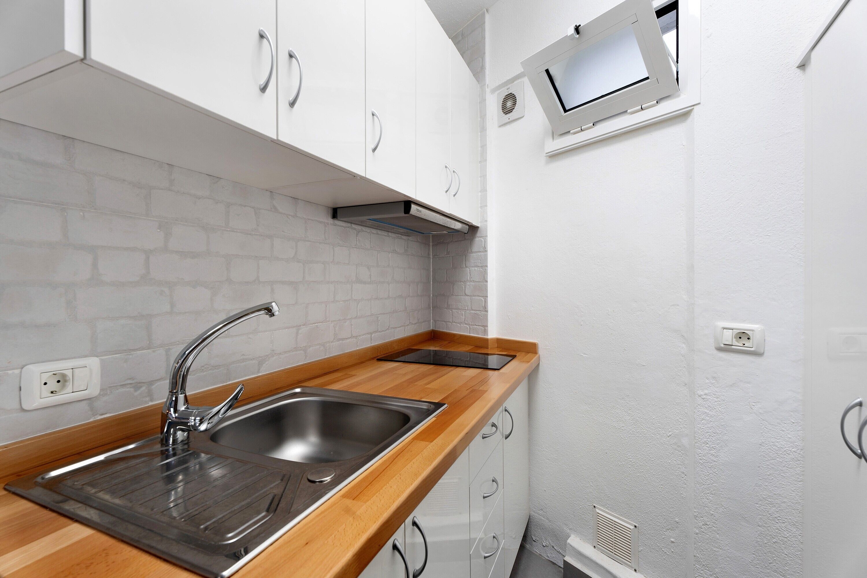 Een keuken met witte kastjes, een houten aanrecht, een roestvrijstalen spoelbak en een raam met een televisie.