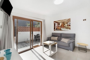 A modern living room with a grey sofa, a small table, a TV, and a sliding glass door leading to an outdoor area.