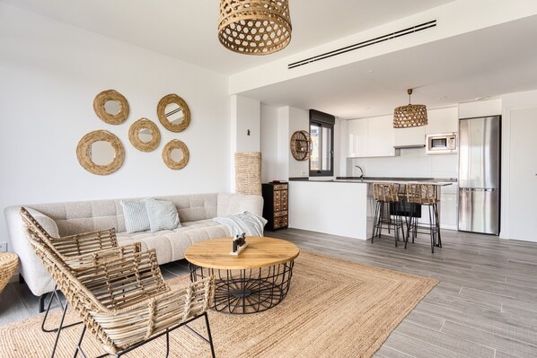 A modern living room with a sofa, coffee table, wicker chairs, and a kitchen area with a dining table. - Apartment 'Izabell' with Mountain View, Shared Pool and Wi-Fi (Finestrat)