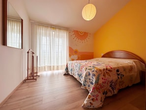A bedroom with a wooden floor, a bed with a floral bedspread, a hanging light fixture, a mirror, and a window with sheer curtains.