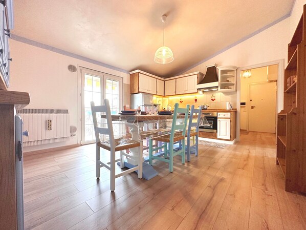 A kitchen with wooden cabinets, a dining table with chairs, a hanging light fixture, and a view of the outside through a glass door.