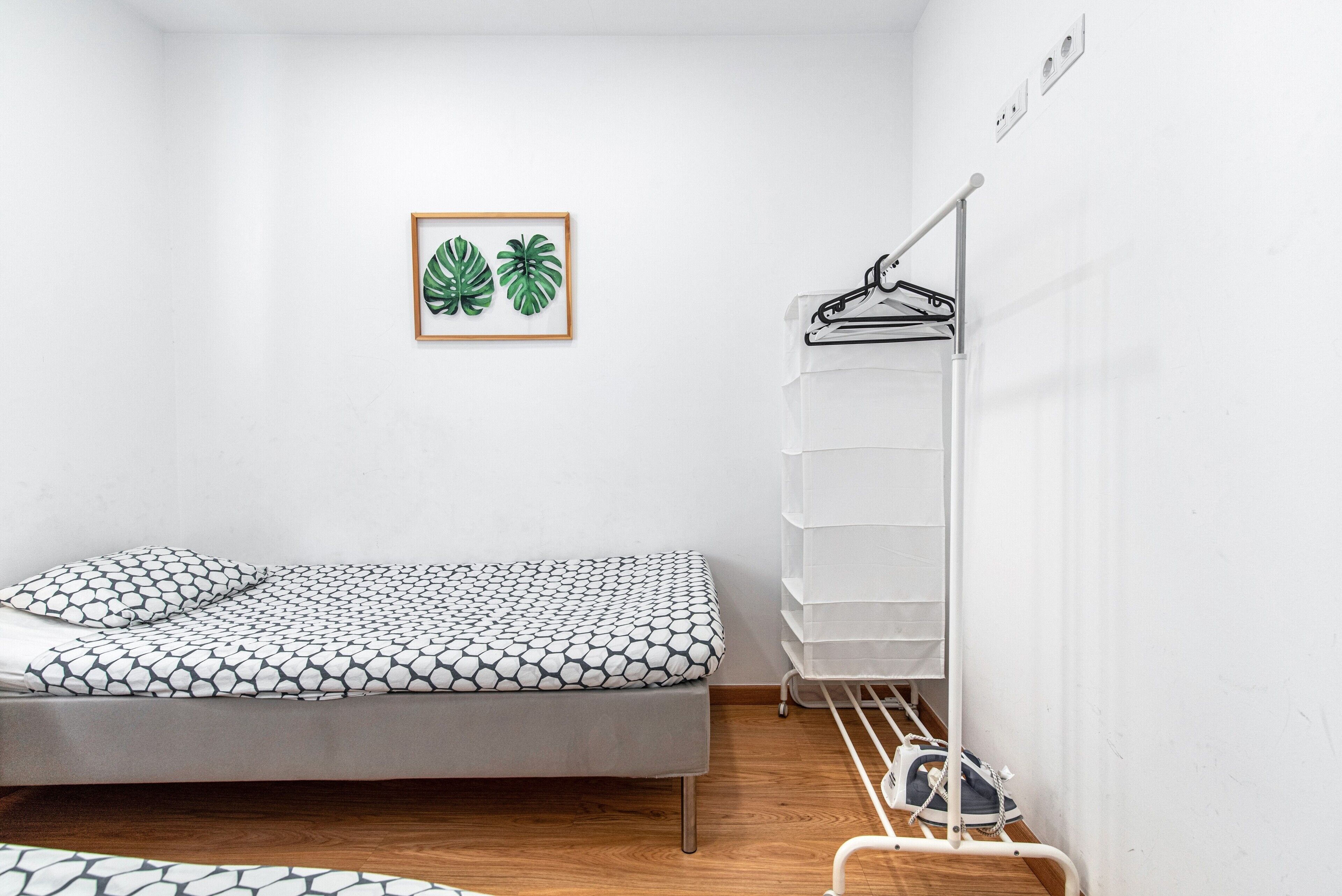 A small, neatly arranged room with a bed, a wall-mounted shelf with hanging items, and a framed picture.
