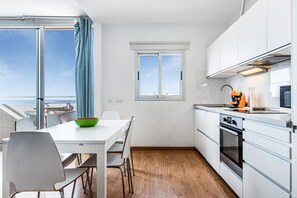 Ett modernt kök med vita skåp, ett trägolv, ett matbord med stolar och en utsikt genom ett stort fönster.