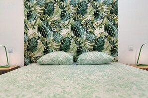 A bed with a patterned bedspread, two pillows, and a wall with a tropical leaf wallpaper.