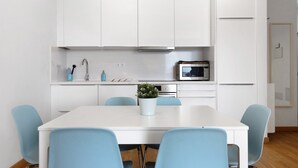 Une cuisine moderne avec des meubles blancs, un four à micro-ondes et un évier. On y trouve une table à manger avec des chaises bleues et une plante en pot.