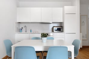 Una cocina moderna con armarios blancos, un microondas y un lavabo. Hay una mesa de comedor con sillas azules y una planta en maceta encima.