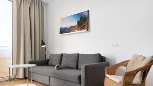 Une pièce avec un canapé gris, un fauteuil en osier, une petite table et un tableau représentant un paysage de montagne.