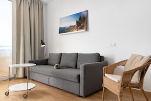 Una habitación con un sofá gris, un sillón de mimbre, una mesita y un cuadro de un paisaje montañoso.
