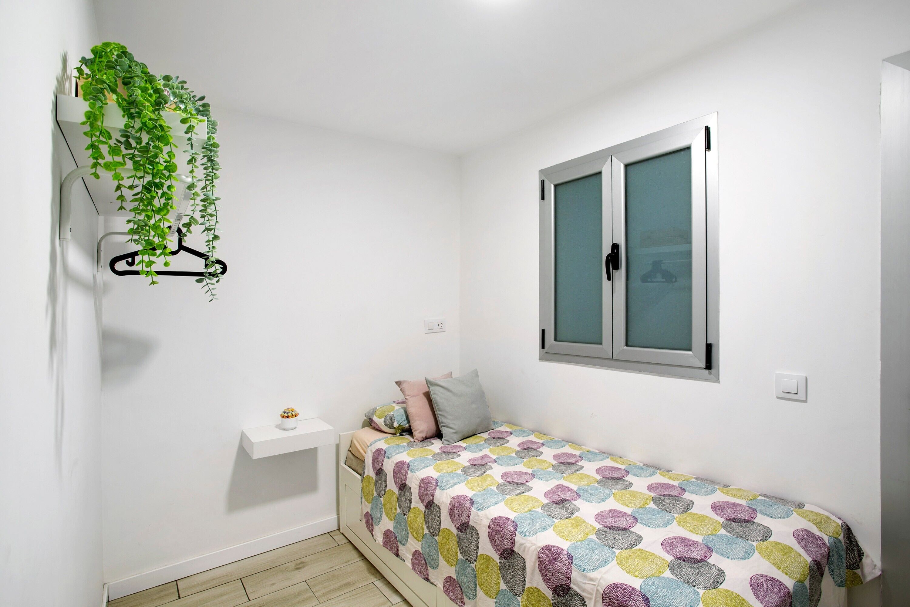 Een kleine, witte kamer met een bed, een raam, een hangende plant en een klein wit plankje.