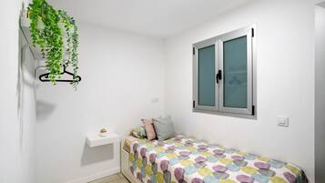 Een kleine, witte kamer met een bed, een raam, een hangende plant en een klein wit plankje.
