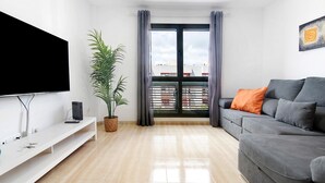 A modern living room with a flat-screen TV, a grey sofa, a potted plant, and a large window with curtains.